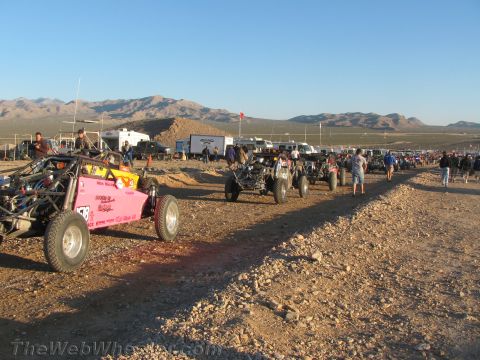 1990 Ford Bronco - pink off-road buggy and other race buggies line up on a gravel staging road in the desert with mountains and trailers behind.