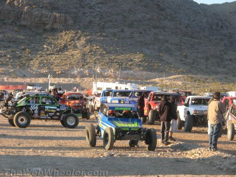 1990 Ford Bronco - Green number 556, a blue buggy and other off-road race cars are staged on gravel at a desert event with spectators nearby.