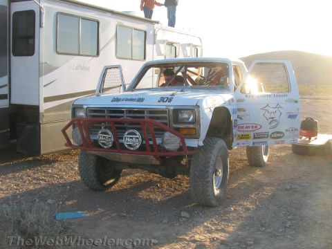 1990 Ford Bronco - white off-road race truck numbered 306 with red Hella-light bumper sits beside a motorhome in low sun at a desert pit.