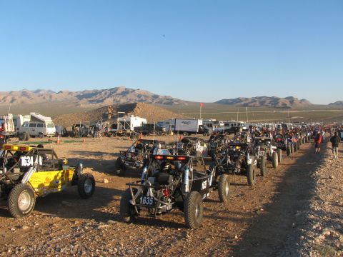 1990 Ford Bronco - Numerous off-road race buggies, including numbers 1634 and 1635, line up in rows on gravel at a crowded desert race start.