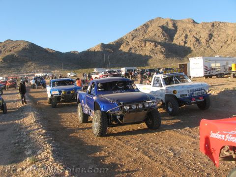 1990 Ford Bronco - blue off-road race truck and a white truck sit among other race vehicles on dirt at a desert event with mountains behind.