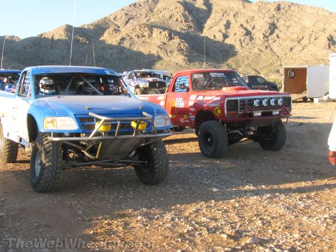 1990 Ford Bronco - blue off-road race truck sits next to a red lifted truck numbered 808 with KC light covers at a desert staging area.
