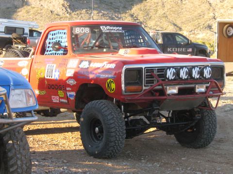 1990 Ford Bronco - red lifted off-road race truck numbered 808 with KC light covers and Mopar Performance lettering sits at a desert pit.