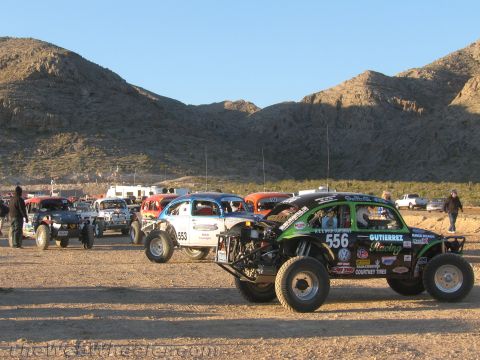 1990 Ford Bronco - green off-road buggy numbered 556 with VW branding and other race buggies are staged on dirt at a desert event near mountains.
