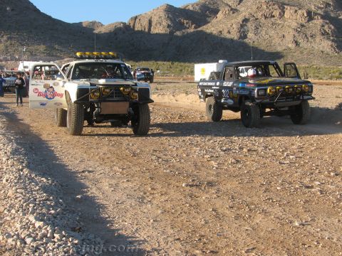 1990 Ford Bronco - white truck and a tan truck numbered 302 with light bars drive along a wide dirt road in the desert.