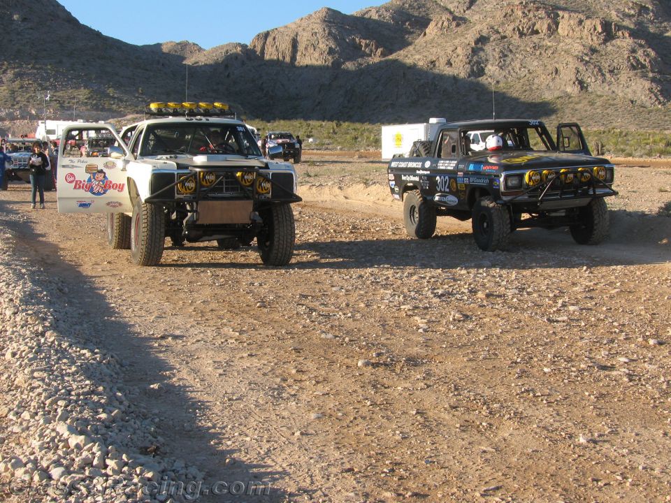 Two off-road race trucks driving on a dirt road through the desert