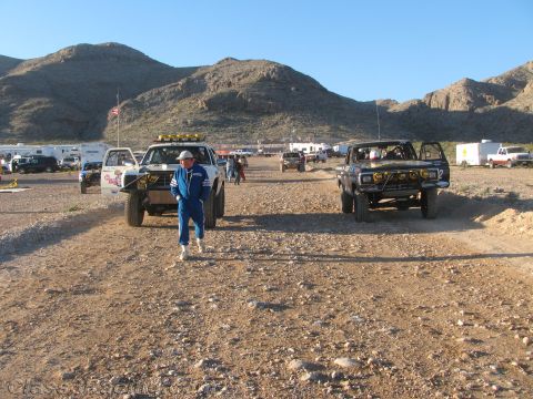 1990 Ford Bronco - person in a blue jumpsuit walks down a dirt road between two parked off-road race trucks at a desert event.