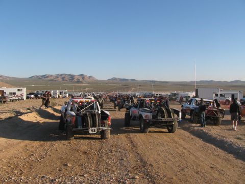 1990 Ford Bronco - long row of off-road race trucks and buggies stage on a desert dirt course at dawn, mountains and chase rigs in the distance.