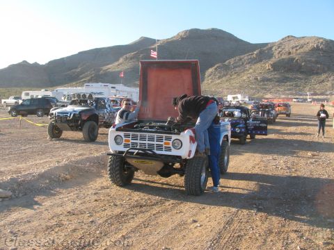 1990 Ford Bronco - Crew members lean into the open engine bay of a white vintage Ford Bronco off-road racer staged in the desert.