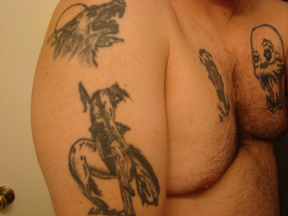 Tattoos of a wolf, cowboy on horse, and skull on a person's torso and arm