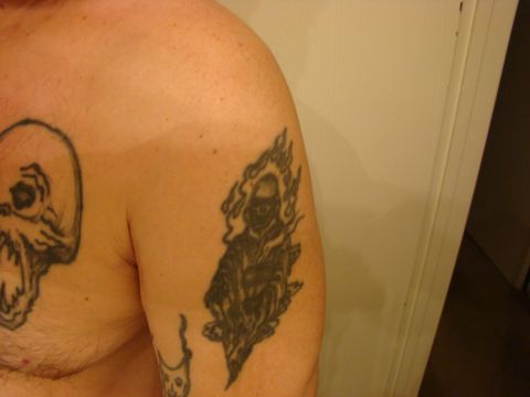 1989 Ford Bronco - Close-up of a person's bare shoulder and chest showing black-and-grey tattoos, including a skull on the chest and a flaming skeleton rider…