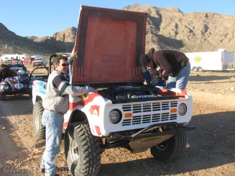 1990 Ford Bronco - Two crew members service the open engine of a white early Bronco off-road racer while a man stands by its raised hood.