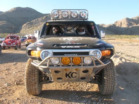 1990 Ford Bronco - Front view of a black Toyota FJ Cruiser off-road racer with a roof light bar and KC-branded bumper in the desert.