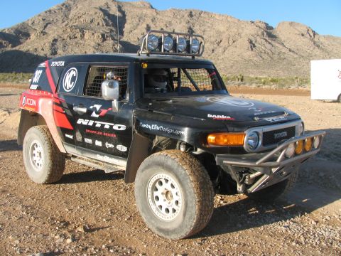 1990 Ford Bronco - black-and-red Toyota FJ Cruiser off-road race truck with Nitto and KC graphics and roof lights, parked on desert ground.