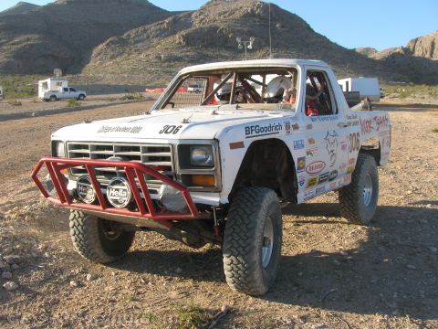1990 Ford Bronco - white number 306 Ford off-road race pickup with a red tube bumper and roof lights sits on rocky desert ground.