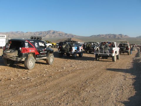 1990 Ford Bronco - Off-road race trucks including a black FJ Cruiser line up along a desert dirt trail with mountains in the distance.