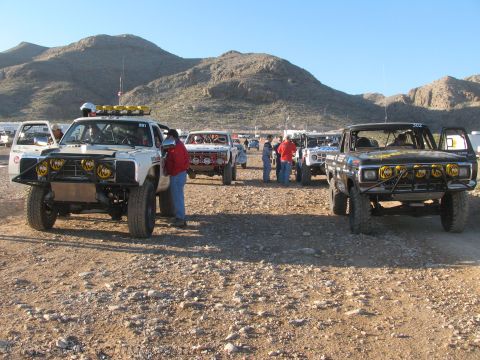 1990 Ford Bronco - Several vintage off-road race trucks with roof light bars sit among standing crew and spectators on rocky desert ground.