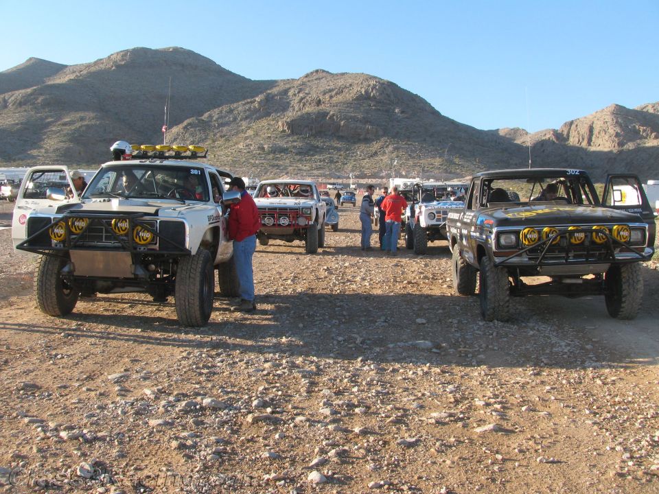 Off-road race trucks parked among spectators in rocky desert lot