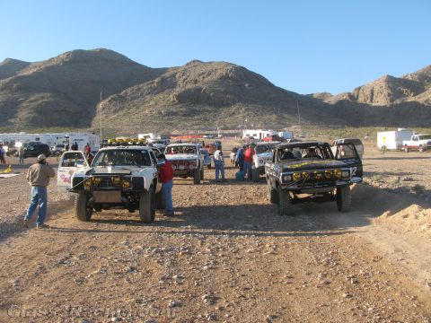 1990 Ford Bronco - Vintage off-road race trucks and crew gather on a rocky desert flat with chase trailers and mountains behind.