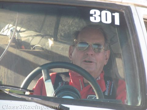 1990 Ford Bronco - man in aviator sunglasses and a red racing suit sits at the wheel inside a race truck marked number 301.