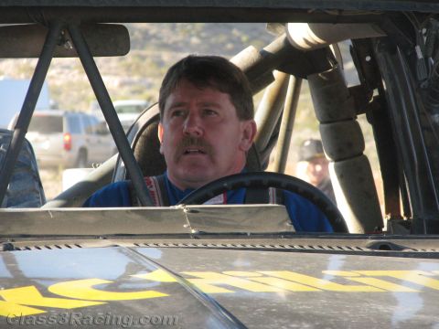 1990 Ford Bronco - mustached man in a blue racing suit sits behind the wheel inside the roll-caged cab of an off-road race truck.