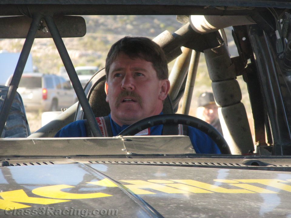Mustached man in blue suit seated inside caged off-road race truck