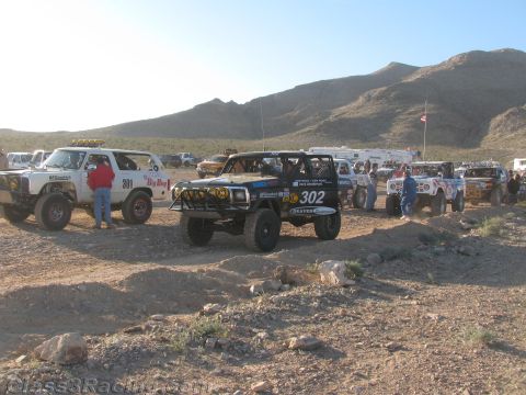 1990 Ford Bronco - black number 302 off-road race truck sits among other staged race trucks and crew on a rocky desert flat.