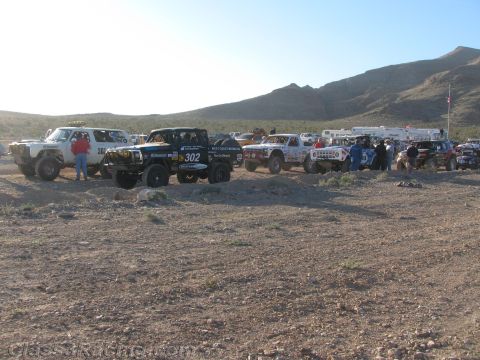 1990 Ford Bronco - line of off-road race trucks led by black number 302 stages across a rocky desert flat with mountains behind.