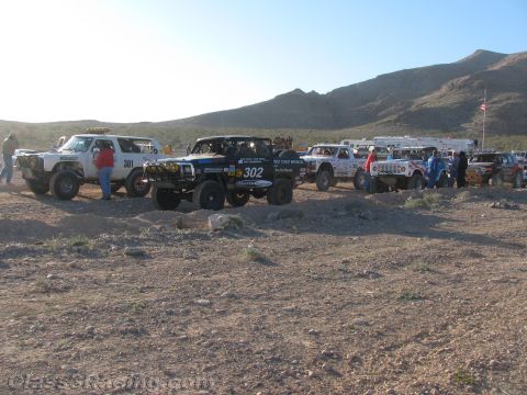 1990 Ford Bronco - Off-road race trucks including white number 301 and black number 302 stage on a rocky desert flat at dawn.