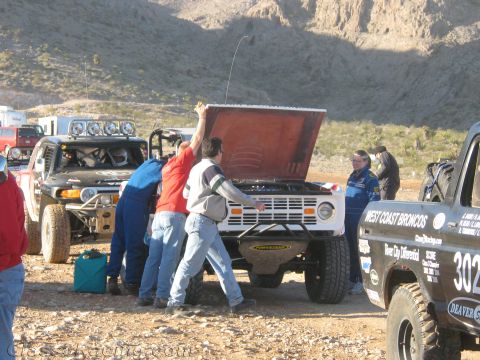 1990 Ford Bronco - Several crew members service a white early Bronco with its hood up beside a West Coast Broncos number 302 race truck.
