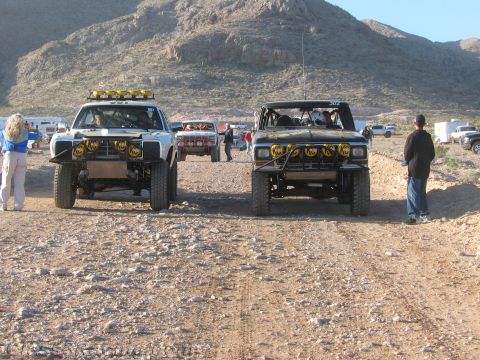 1990 Ford Bronco - white and a black off-road race truck park nose-out on a desert dirt trail with crew standing alongside.