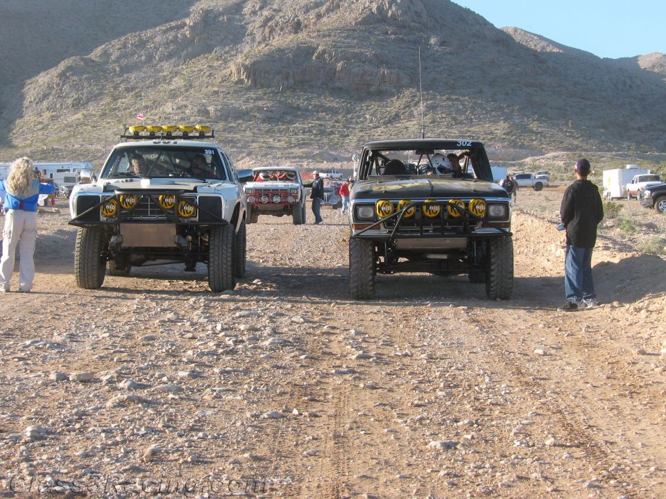 Two off-road race trucks parked nose-out on desert trail with crew