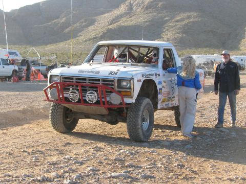 1990 Ford Bronco - white number 306 Ford off-road race truck with red tube bumper sits in the desert as two people stand beside it.