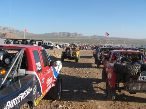 1990 Ford Bronco - Dense rows of colorful off-road race trucks and buggies stage across a desert flat with flags and a large crowd.