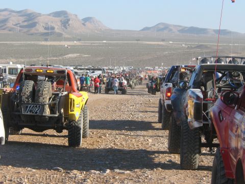 1990 Ford Bronco - Two columns of off-road race trucks and buggies stage along a wide desert course as crews and spectators gather.