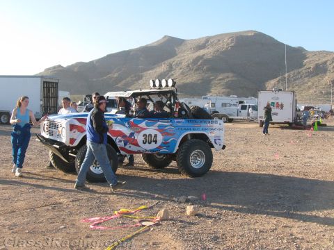 1990 Ford Bronco - red-white-and-blue early Ford Bronco number 304 off-road racer sits in a desert pit area as crew walk nearby.