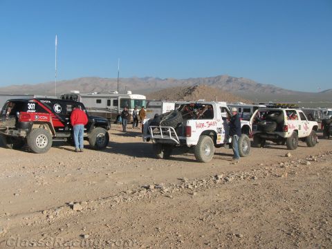 1990 Ford Bronco - Off-road race trucks including number 307 stage near RVs and trailers across an open desert flat under clear sky.