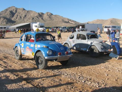 1990 Ford Bronco - Two desert-race-prepped Volkswagen Beetles, a blue car numbered 103 and a silver car numbered 1107, sit on dirt near support trailers and m…