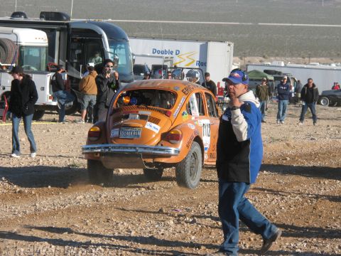 1990 Ford Bronco - orange off-road-prepped Volkswagen Beetle numbered 2112 is seen from behind in a dusty staging area as a man in a blue jacket walks past.