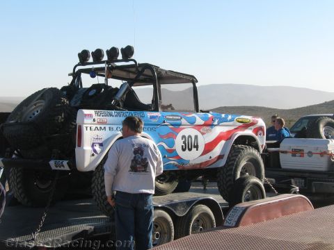 1990 Ford Bronco - American-flag-themed off-road buggy numbered 304 with roof light bar sits chained on a tandem-axle trailer while a man stands at its side.