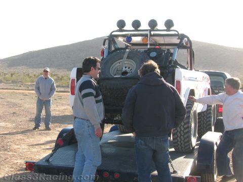 1990 Ford Bronco - Three men gather behind an off-road race vehicle loaded on a flatbed trailer, with a spare tire and roof light bar visible in the desert.