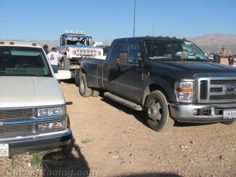 1990 Ford Bronco - black Ford dual-rear-wheel pickup and a white pickup sit in a dusty pit area, with a race buggy on a trailer in the background.