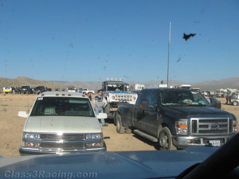 1990 Ford Bronco - black Ford dual-rear-wheel pickup tows a trailer next to a silver van numbered 306 amid a crowded desert race pit under blue sky.
