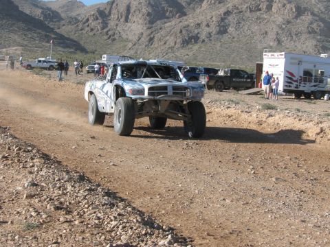 1990 Ford Bronco - silver-and-blue off-road race truck speeds along a dirt course kicking up dust, with spectators and trailers along the edge.