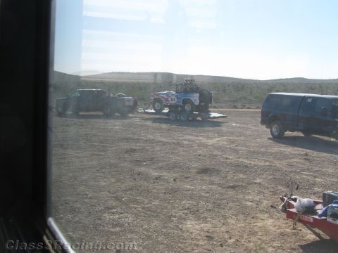 1990 Ford Bronco - Seen through a tinted window, a race vehicle sits on a trailer beside trucks and a dark van across a dusty desert pit.