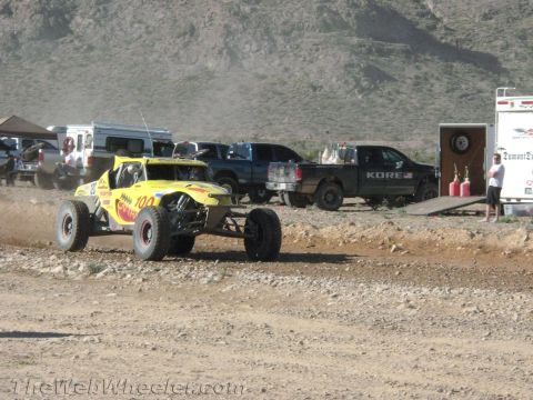 1990 Ford Bronco - yellow open-cockpit off-road buggy numbered 700 rounds a dirt course with support trucks, a KORE pickup and trailers in the background.