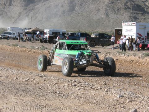 1990 Ford Bronco - green-and-white open-wheel off-road buggy carrying a Goodyear banner races across a dirt course past spectators and support trailers.