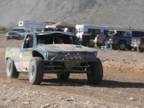 1990 Ford Bronco - dark, mud-streaked off-road race truck numbered 44 corners on a dirt course with support trucks and trailers lined up behind.