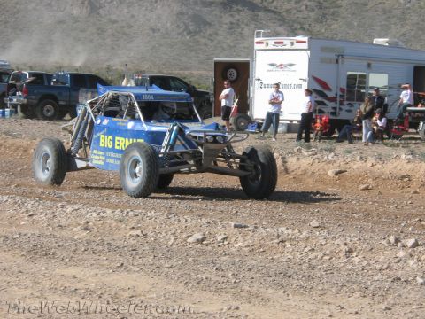 1990 Ford Bronco - blue open-frame off-road buggy lettered 'BIG ERNIE' drives a dirt course as spectators sit by trailers in the background.