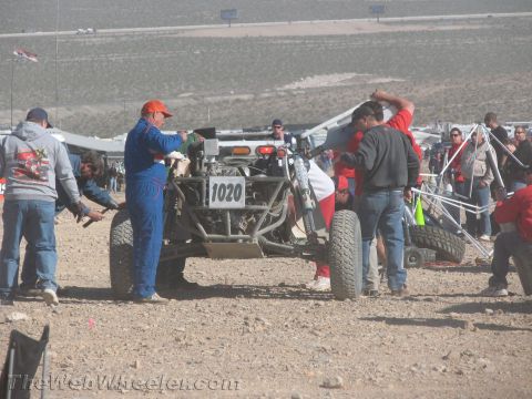 1990 Ford Bronco - crew clusters around an open-frame off-road buggy numbered 1020 being serviced in a busy desert pit dotted with canopies.